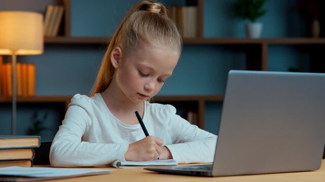 Caucasian concentrated little child pupil student school girl daughter kid writing notes e-learning studying distant at home remote education write homework looking in laptop screen study in evening