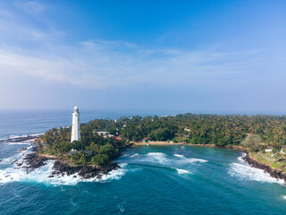 Aerial drone photo of Dondra Head Lighthouse viewed from the sea on a bright sunny day, Sri Lanka. Tall white lighthouse standing on the southernmost point of the island, surrounded by deep blue ocean