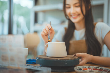 Happy young Asian woman painting a white ceramic mug with a brush in a pottery studio for creative hobby and handmade craft concept