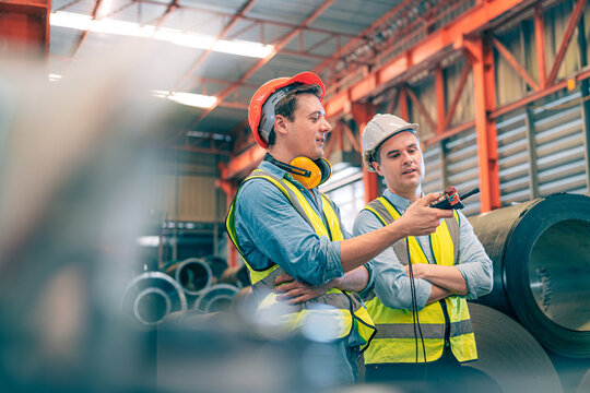 Caucasian industrial engineer men controlling overhead crane with remote in factory. Worker partner wear safety helmet vest lifting heavy metal coil material. Manufacturing hoist machine logistics - Powered by Adobe