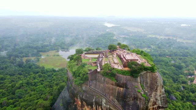 Early morning aerial drone footage of Sigiriya Rock Fortress after rain, Sri Lanka. Low clouds and mist drifting above lush tropical jungle, with wet surfaces reflecting soft morning light.Atmospheric