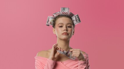 A woman in a pink blouse, with curlers shaped like banknotes in her hair against a pink background, tears up a banknote. A humorous concept of wealth.