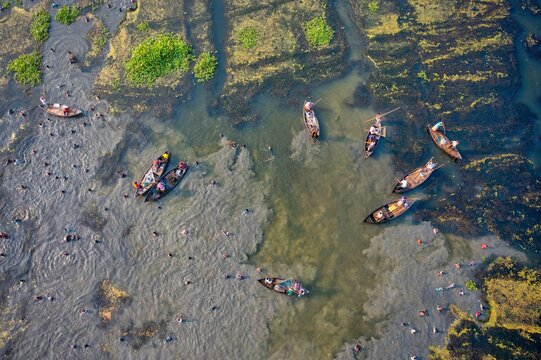 Pabna, Bangladesh - 27 November 2021: Aerial view of boats scattered across the muddy riverbed, their dark forms contrasting with the shimmering water and patches of bright green vegetation.
