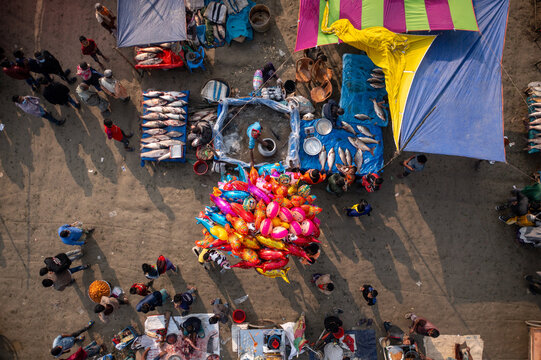 Bogura, Bangladesh - 10 February 2021: Aerial view of a bustling market scene unfolds, vibrant canopies casting shadows over vendors and shoppers amidst colorful balloons and stacks of fresh fish.