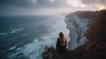 A person sits on the edge of a cliff and looks at the ocean waves beneath. The sky is cloudy and the trees in the background show autumn colors.