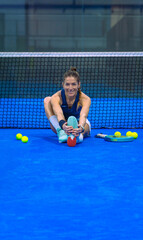 Female padel player, smiling and looking at the camera, stretching her hamstrings on a blue padel court in front of the net.