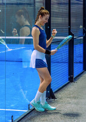 A happy padel player celebrates a victory, looking at her padel racket with her fist raised in a gesture of satisfaction next to the glass of the indoor padel court.
