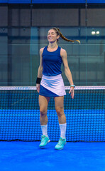 A female padel player in sportswear and a skirt jumps for joy, her hair in a ponytail, dancing during her warm-up on an indoor sports court.