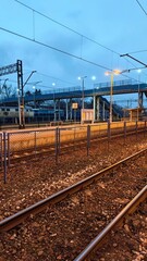 Railway tracks leading through an empty train station platform at dusk with overhead lines and pedestrian bridge. The image represents travel, transportation infrastructure, and urban transition
