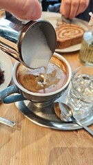Cup of coffee with fresh milk foam being poured from a metal pitcher on a wooden cafe table. The image represents coffee ritual, craftsmanship, and cozy social moment