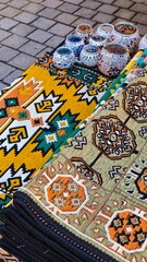 Colorful traditional woven rugs with ethnic geometric patterns displayed at a street market with mosaic glass candle holders on top. The image represents cultural heritage craftsmanship souvenir