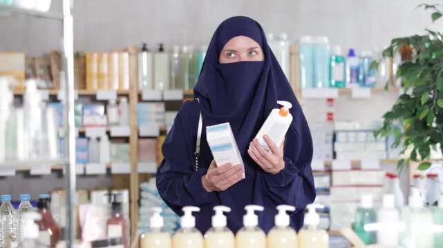 Young Muslim woman in traditional blue niqab(hijab) selecting body cream in pharmacy, thoughtfully comparing product options and reading labels before making decision 