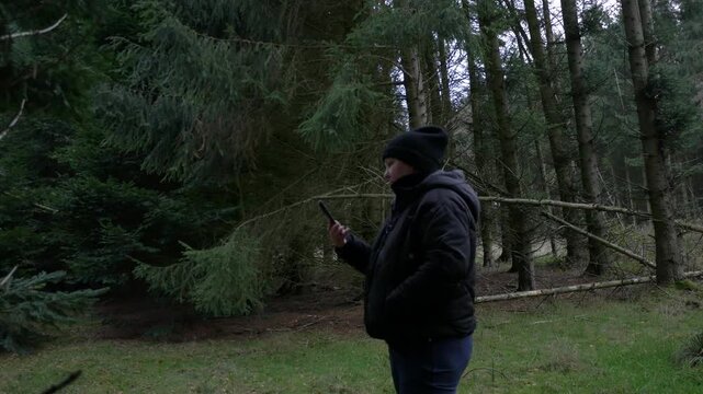 Woman in a black jacket and hat stands in a dark, dense forest, looking down at a smartphone in her hands to check the hiking route or navigation app.