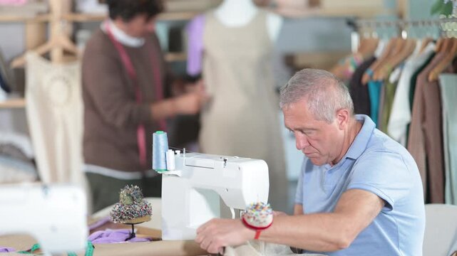 Experienced tailor sews clothes against the background of his assistant, who checks a sample of the dress on a mannequin. Male tailors work on ordering clothes in the workshop. High quality 4k footage