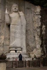 A person meditates near a towering stone Buddha statue carved into a cliff.