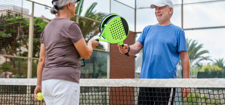 Senior couple playing paddle tennis or padel, outdoors in padel court enjoying and having fun together. Mature woman and old man high five after finish game