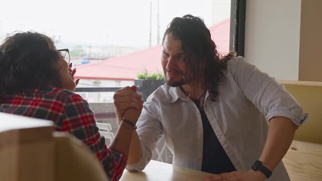 Playful LGBTQ friends arm wrestling at a cafe table indoors