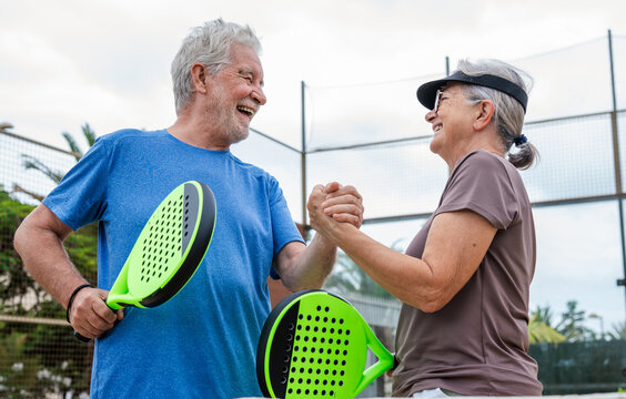 Senior couple playing paddle tennis or padel, outdoors in padel court enjoying and having fun together. Mature woman and old man high five after finish game