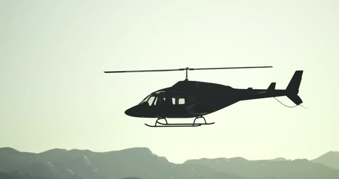 A helicopter is seen flying over a mountain range during sunset. The silhouette contrasts against the colorful sky, highlighting the serene yet adventurous atmosphere of the moment.