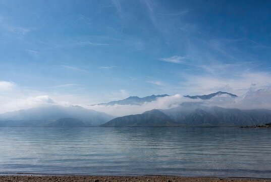 Majestic mountains shrouded in mist and clouds rise above a calm lake under a clear blue sky - Powered by Adobe