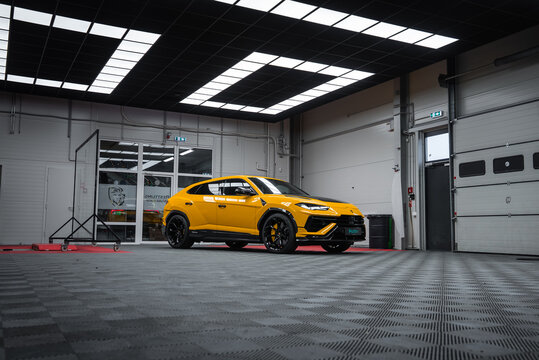 A yellow Lamborghini Urus Performante is parked indoors with black wheels, carbon fiber accents, roll up doors, glass entry signage, and ceiling light panels on a gray floor.