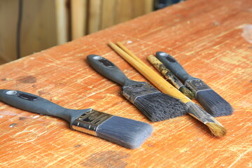 Gebrauchte Malpinsel auf zerkratztem Holzbrett: drei breite Synthetikpinsel und zwei feine Naturhaarpinsel mit Farb Sprenkeln und sichtbarer Abnutzung, Atelier-Atmosph&auml;re.