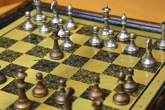 Metallisches Schachspiel auf gelb-schwarzem Brett, stilvoll arrangiert auf einem Holztisch. Silberne und bronzene Figuren in Startposition &ndash; ein Hauch von Eleganz und Strategie.