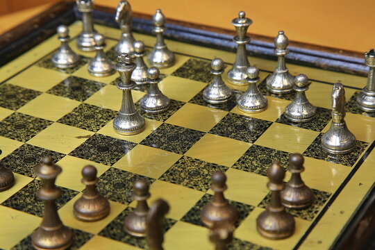 Metallisches Schachspiel auf gelb-schwarzem Brett, stilvoll arrangiert auf einem Holztisch. Silberne und bronzene Figuren in Startposition &ndash; ein Hauch von Eleganz und Strategie.