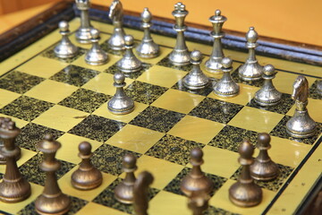 Metallisches Schachspiel auf gelb-schwarzem Brett, stilvoll arrangiert auf einem Holztisch. Silberne und bronzene Figuren in Startposition &ndash; ein Hauch von Eleganz und Strategie.