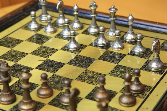 Metallisches Schachspiel auf gelb-schwarzem Brett, stilvoll arrangiert auf einem Holztisch. Silberne und bronzene Figuren in Startposition &ndash; ein Hauch von Eleganz und Strategie.