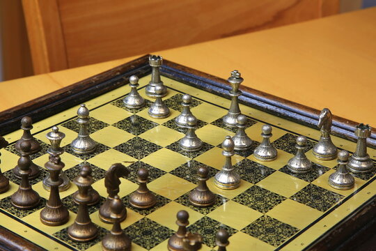 Metallisches Schachspiel auf gelb-schwarzem Brett, stilvoll arrangiert auf einem Holztisch. Silberne und bronzene Figuren in Startposition &ndash; ein Hauch von Eleganz und Strategie.