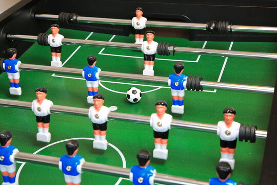 Kicker-Nahaufnahme: Ball liegt zentral auf dem gr&uuml;nen Spielfeld, flankiert von blauen und wei&szlig;en Kunststoffspielern auf Metallstangen; ein dynamischer Moment im Tischfu&szlig;ballspiel.