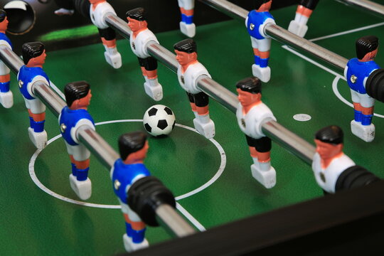 Kicker-Nahaufnahme: Ball liegt zentral auf dem gr&uuml;nen Spielfeld, flankiert von blauen und wei&szlig;en Kunststoffspielern auf Metallstangen; ein dynamischer Moment im Tischfu&szlig;ballspiel.