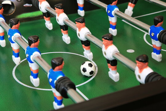 Kicker-Nahaufnahme: Ball liegt zentral auf dem gr&uuml;nen Spielfeld, flankiert von blauen und wei&szlig;en Kunststoffspielern auf Metallstangen; ein dynamischer Moment im Tischfu&szlig;ballspiel.