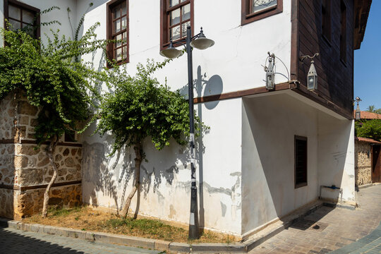 Corner of traditional Ottoman house with wooden bay windows and tile roof in old town Kaleici. Historic lanterns, cobblestone street, and greenery under blue sky. Mediterranean, Antalya, Turkey.

