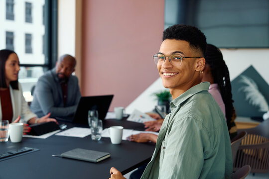 Confident young businessman smiling at camera while attending team meeting in boardroom at office