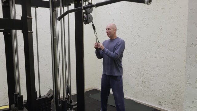 Athletic bald man in blue sweatshirt executing standing cable triceps pushdown exercise with rope attachment on black weight machine in bright modern home gym.