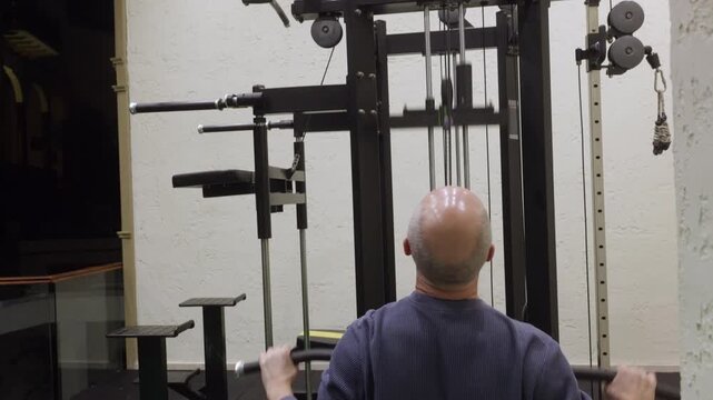 Rear view of athletic bald man in blue ribbed sweatshirt and navy pants performing wide grip lat pulldown exercise on black cable machine in modern home gym with white walls.