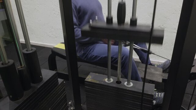 Athletic bald man in blue ribbed sweatshirt and navy pants sitting on yellow bench performing seated cable row back exercise on black multi station machine with green weight markers.