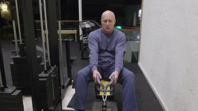 Focused bald man in blue sweatshirt and navy pants sitting on yellow workout bench holding cable handles on black multi station gym equipment in contemporary fitness room.