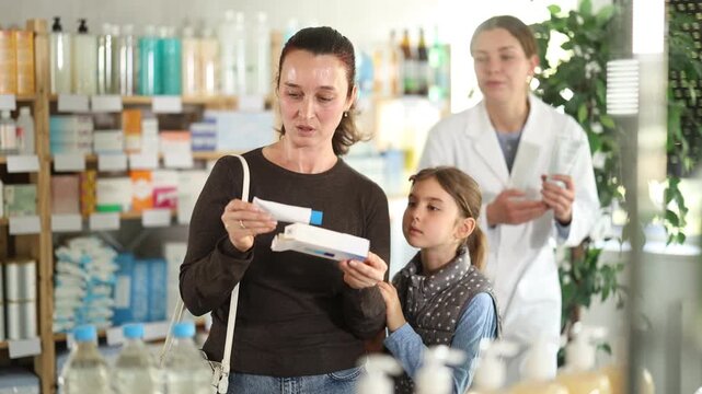 Woman and daughter are standing in a pharmacy and choosing a medical ointment for skin irritation and psoriasis against the background of a pharmacist. Mom and child shopping at a pharmacy in autumn