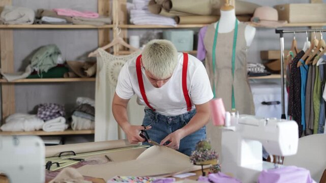 Stylish young guy seamstress cutting pieces of paper with scissors according to drawn template for pattern during dress design at workplace in modern sewing studio 