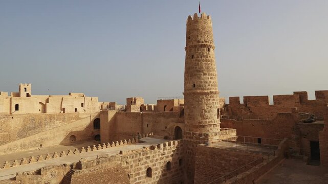 Inner courtyard of Ribat Harthama in Monastir showcasing traditional historic stone architecture and tall cylindrical watchtower bathed in golden sunlight under clear Tunisian sky. High quality 4k