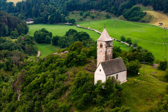 Chiesa di Santa Verena on a gentle hilltop with fields below, aerial view. Dolomites landscape with agricultural parcels
