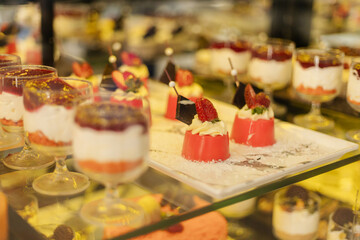 Different types of desserts are arranged neatly on a glass shelf in a bakery. People walk by and look at the tasty treats, enjoying the atmosphere of the market