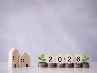 Miniature house, Wooden blocks with number 2026 and plants growing up on stack of coins. The concept of saving money for house, Property investment, House mortgage, Real estate in year 2026.