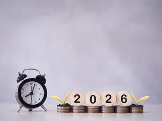 Black alarm clock, Wooden blocks with the number 2026 and plants growing up on stack of coins. The concept of saving money for manage time to success business in year 2026