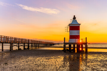 Vecchio Faro lighthouse in Lignano Sabbiadoro at golden sunset, historic red and white beacon by wooden pier reflecting in shallow sea, iconic Adriatic coast landmark in Italy, travel destination.