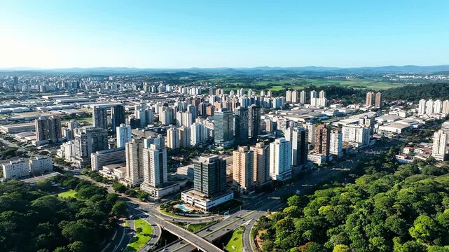 Panoramic aerial drone flight over a modern city skyline with lush green trees and clear blue sky