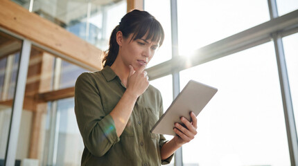 Professional woman standing in a bright modern office, thoughtfully reading or analyzing something on a digital tablet device.